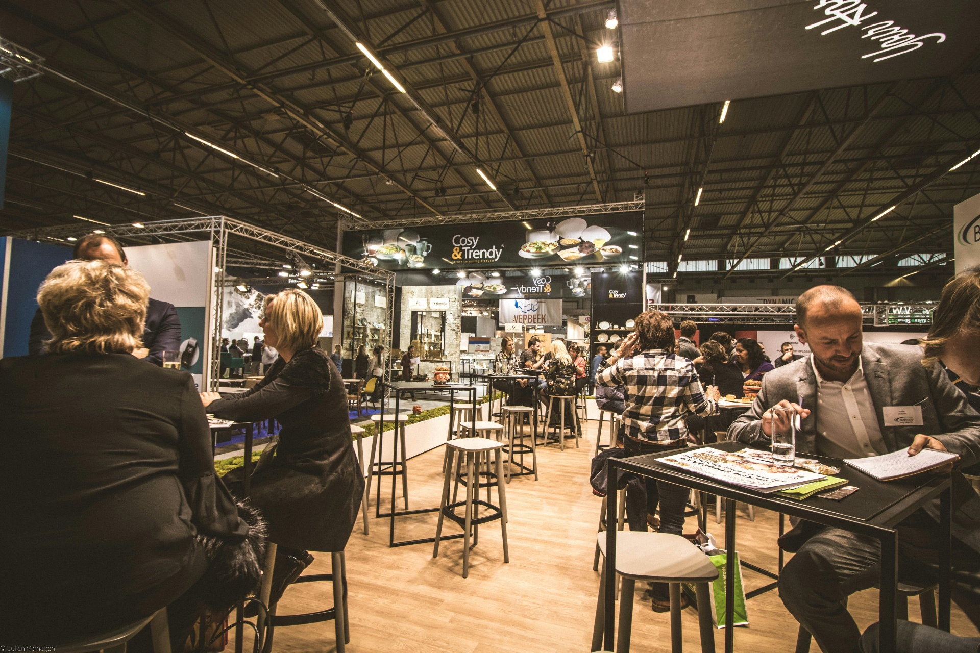 Wide view of a busy trade expo with attendees networking at supplier booths under industrial roof trusses