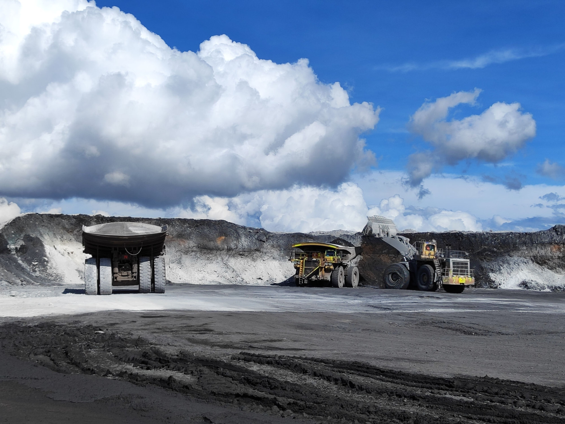 Haul trucks and a wheel loader working an open-cut Australian mine under a bright cloudy sky