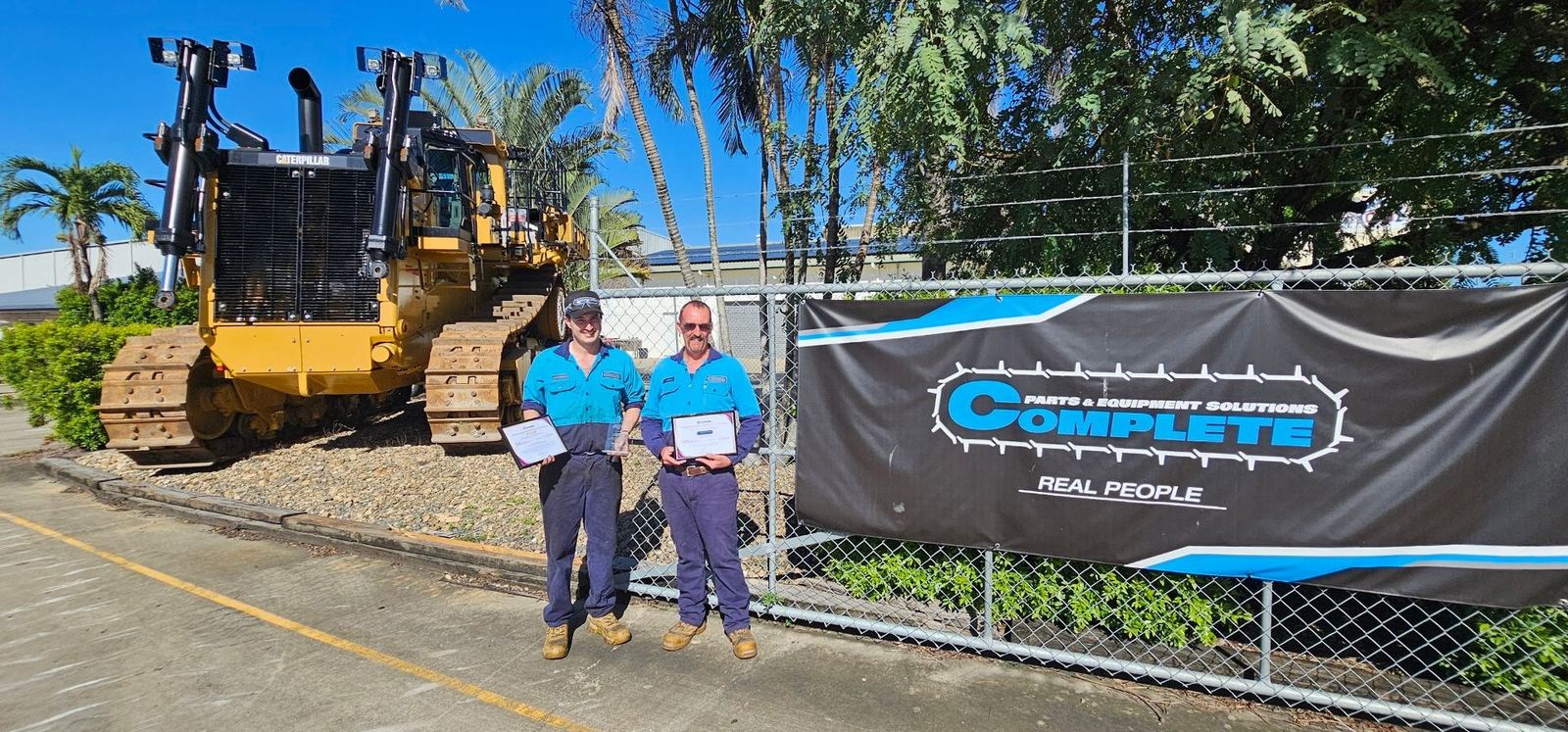 Mackenzie and colleague holding awards at Complete Parts and Equipment Solutions with CAT machinery in background