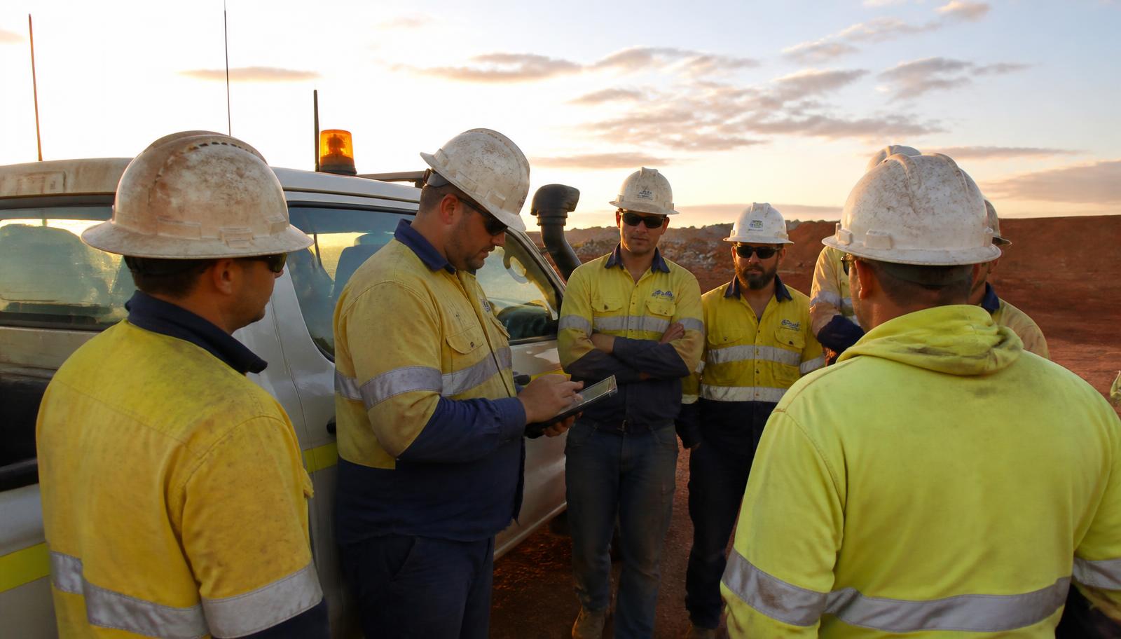 A supervisor marking off a crew list on a rugged tablet at a dawn pre-start meeting beside a light vehicle on a Western Australian mine site