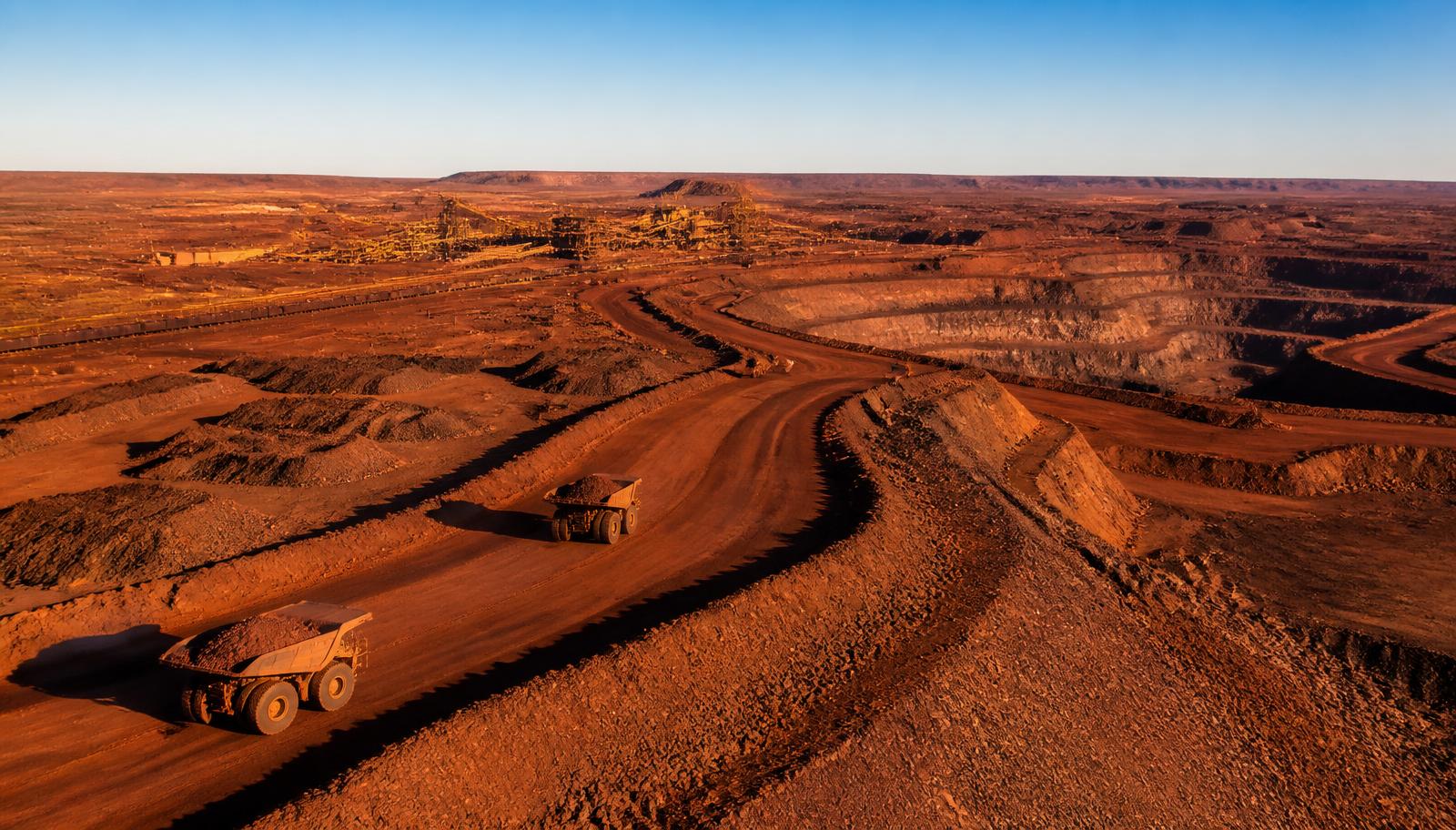 Aerial view of a large Pilbara iron ore mine in Western Australia at golden hour with haul trucks on red earth ramps and a processing plant in the distance